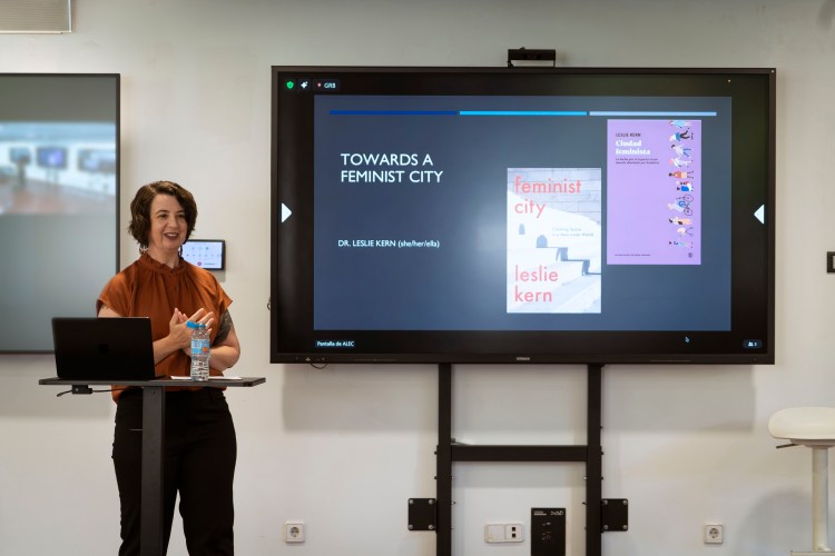Leslie at a lectern in front of a slide show with the slide reading: Towards a Feminist City.