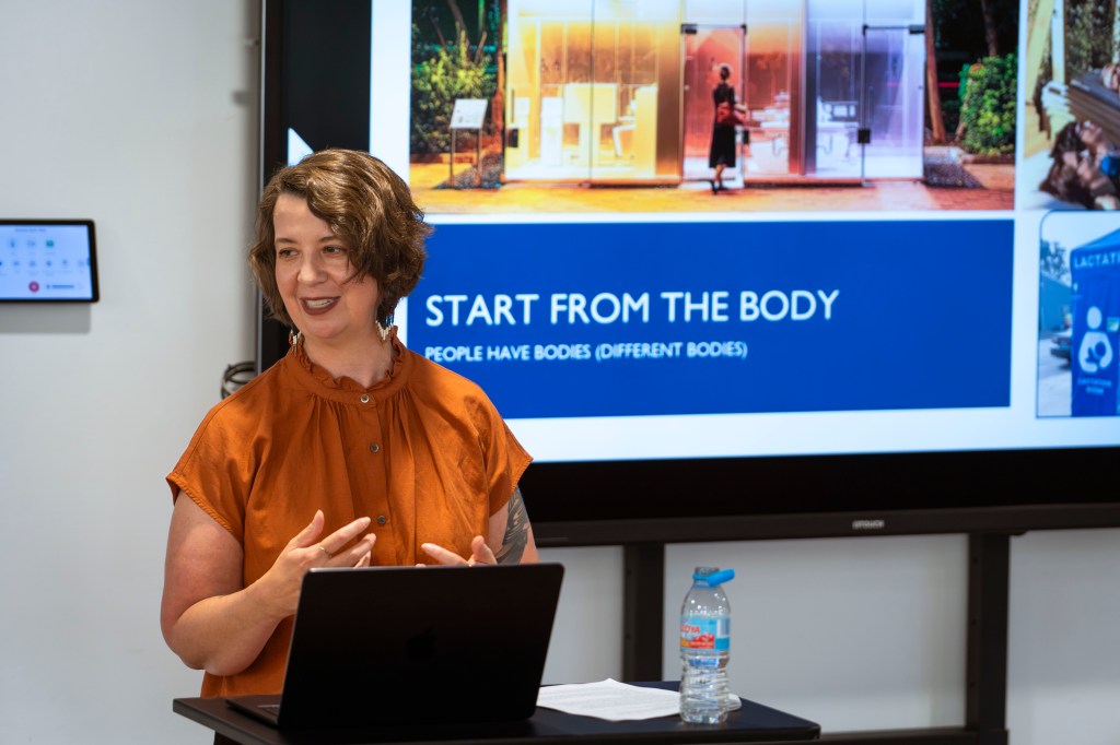 Leslie in an orange shirt standing at a lectern in front of a slide show.