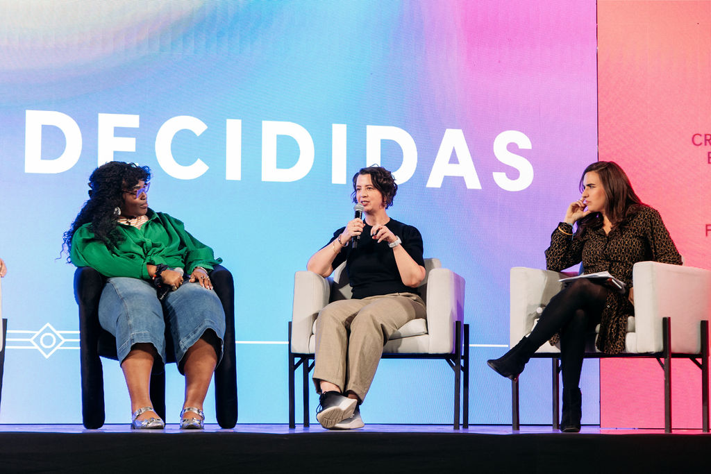 3 women speakers sit in chairs on a stage at Decididas Forum. Leslie is in the middle.